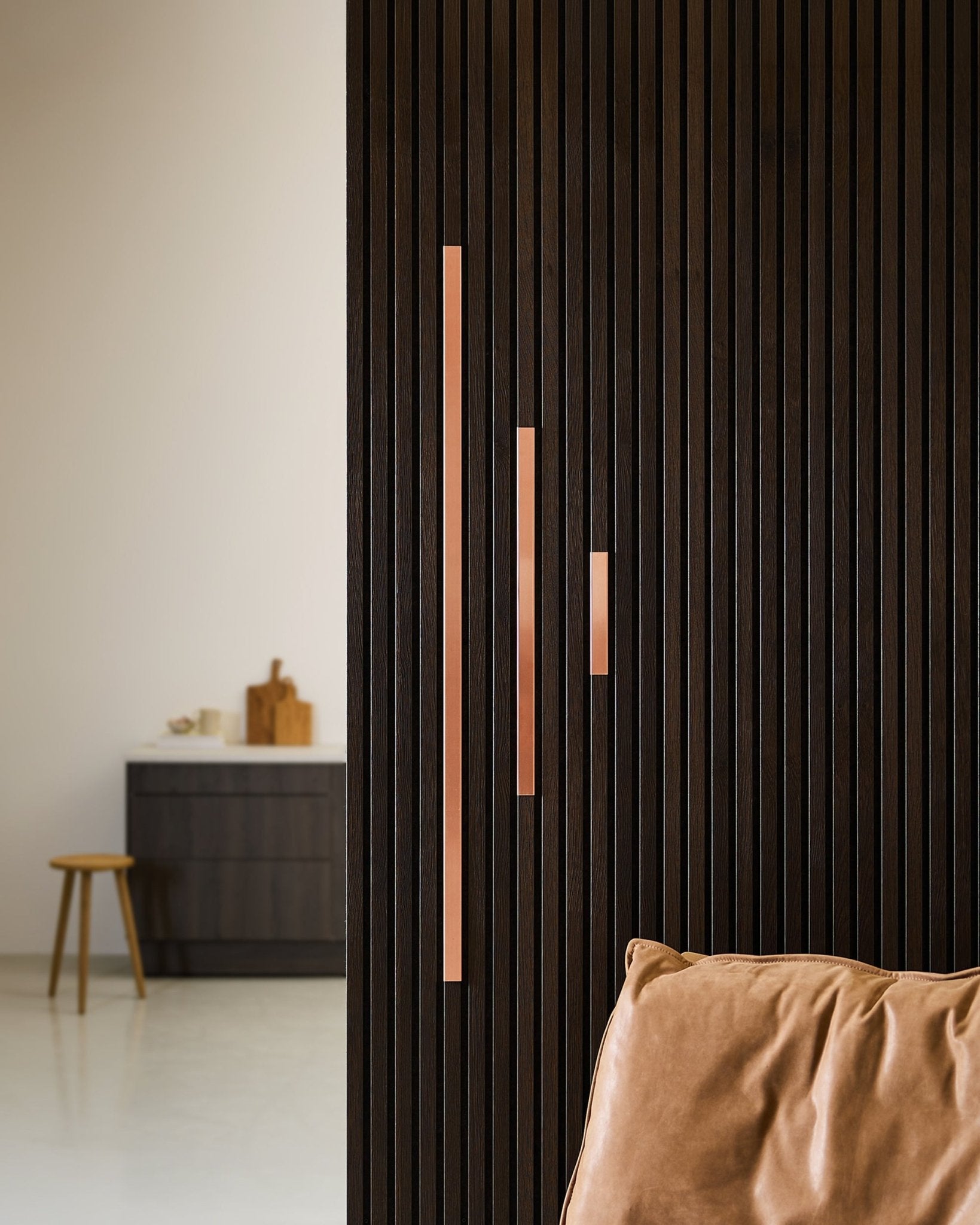 Minimalist space featuring a dark wood acoustic slatted partition with WoodUpp Liva Cover in copper, 60 cm panels with copper handles, tan leather chair, and cabinet stool.